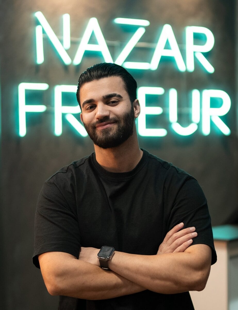 Handsome man with dark hair and beard standing in front of neon sign at Nazar Friseur, modern hair salon in Berlin.